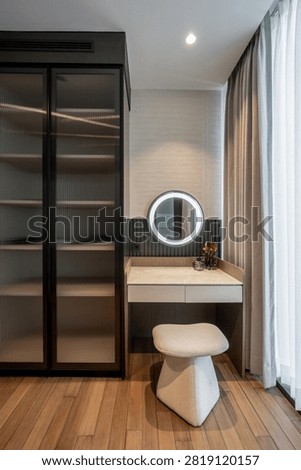 Modern bedroom dressing table interior with illuminated round mirror, cream stool, and fluted glass wardrobe.