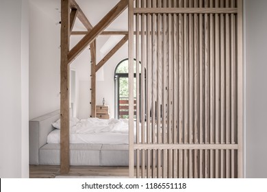 Modern bedroom with white walls, parquet, wooden beams and a lattice partition. There is a gray bed with light linens, doors to a balcony, wood chests with a statuette, hanging filament lamps. - Shutterstock ID 1186551118