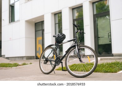 Modern Bicycle Standin On Footplate In City Street On Blurred Background Of Front Door Of Apartment Building, Office Building. Protective Helmet Lies On Seat Of Bike, Outdoors, No People, Nobody.