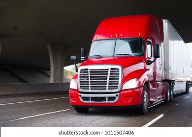 A modern big rig semi truck for long haulage with a high cabin for improving aerodynamic characteristics moves under the bridge transporting a dry van semi trailer with commercial cargo