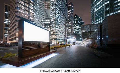 Modern Billboard With A Green Screen On A Busy Street Of The Big City In The Evening. Tall Skyscrapers Glittering With Neon Lights, Road Traffic And Pedestrians Passing By.