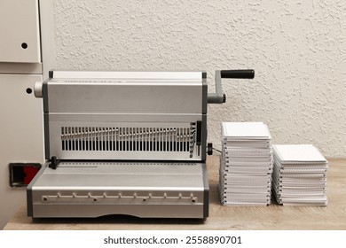 Modern binding machine and stacks of notebooks on wooden table indoors