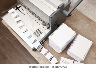 Modern binding machine, stacks of notebooks and double loop wire binding spines on wooden table indoors, above view - Powered by Shutterstock