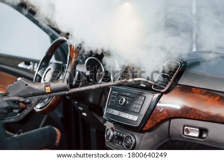 Modern black automobile get cleaned inside by steam equipment by worker inside of car wash station.