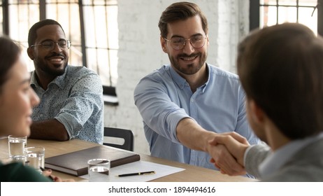 In Modern Board Room Gathered Confident Partners Sit At Desk Businessmen Shake Hands Express Respect Starting Formal Meeting In Office. Closing Deal, Buy Services, Satisfied Client Glad Boss Concept