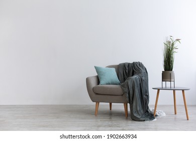 Modern boho cozy corner of home in living room. Gray vintage armchair with blue pillow and blanket, table with green plant in pot, wooden floor, on light wall background, copy space, mockup, nobody