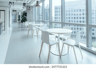 A modern break area in an office, featuring large windows that allow ample natural light to flood the space. The area is furnished with minimalist white tables and chairs. - Powered by Shutterstock