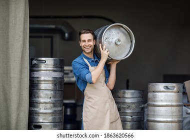 Modern brewery and worker with keg on plant, small business and beer industry. Smiling handsome young strong employee man in apron carries metal barrel on shoulder in warehouse interior, free space - Powered by Shutterstock