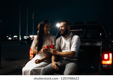 Modern bride and groom is sitting in trunk of car and drink cola. Wife and husband on the boot of car - Powered by Shutterstock