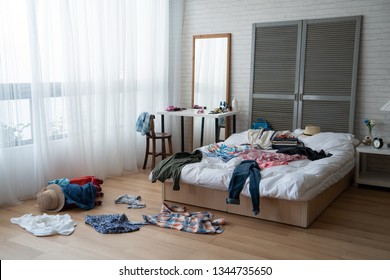 Modern Bright Bedroom With Messy Clothes Scatter On White Bed And Floor. Empty Room With Nobody In Cozy Apartment. Packing Luggage Suitcase For Summer Vacation And Spring Holidays Concept Lifestyle.