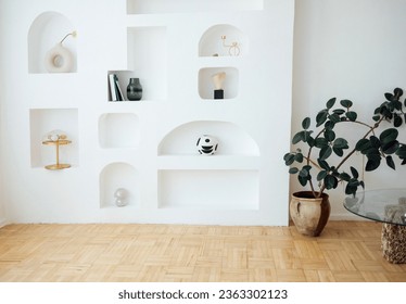 Modern bright interior of living room with white wall and niches. Light parquet, potted plant and coffee table with glass top. Vases, golden color lamp and candlestick, stones, books on shelves.