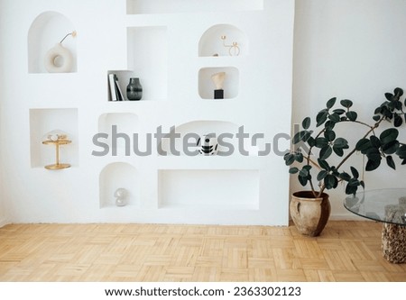 Modern bright interior of living room with white wall and niches. Light parquet, potted plant and coffee table with glass top. Vases, golden color lamp and candlestick, stones, books on shelves.