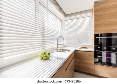 Modern bright kitchen interior with white horizontal window blinds, wooden cabinets with white countertop and household appliances - Shutterstock ID 693495286