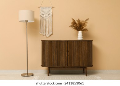 Modern brown chest of drawers, floor lamp and vase with pampas grass near beige wall in room