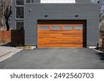 A modern brown faux wooden exterior garage door with four small horizontal glass windows. The modern door is on a luxury dark grey contemporary house with a concrete driveway. 