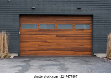 A modern brown faux wooden exterior garage door with four small horizontal glass windows. The modern door is on a luxury dark grey contemporary house with a concrete driveway.  - Shutterstock ID 2249479897