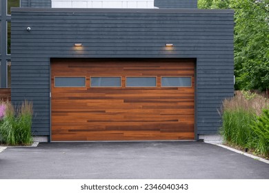 A modern brown faux wooden exterior garage door with four small horizontal glass windows. The modern door is on a luxury dark grey contemporary house with a concrete driveway.  - Powered by Shutterstock