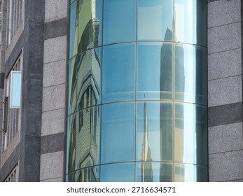 Modern building facade featuring curved glass panels reflecting neighboring skyscrapers. The architecture combines grey stone tiles with blue glass windows in a contemporary urban setting.