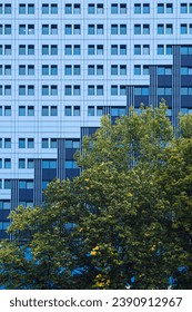 Modern building facade with rhythmic windows patterns against vibrant foliage of green trees. Urban architecture meets nature. Urban planning, architecture, green cities, environmental sustainability