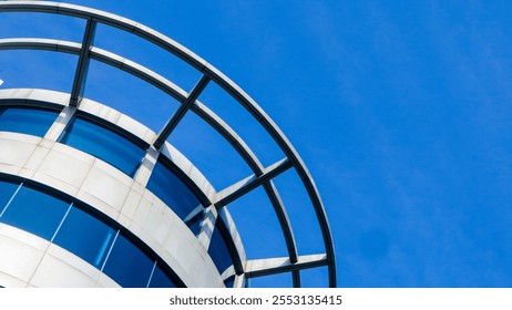 a modern building's curved facade against a clear blue sky. The building features large glass windows and a distinctive metal frame, creating a striking architectural detail. - Powered by Shutterstock