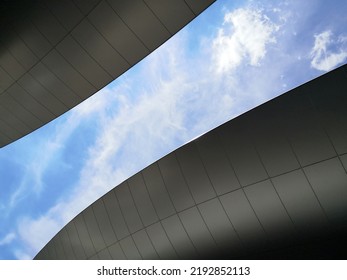 Modern Buildings, View From Below. Abstract Architecture And Material Design With Curvy Hitech Roof. Minimal Exterior Background With Curve Structure. Geometric Composition Of Curves Against Blue Sky.