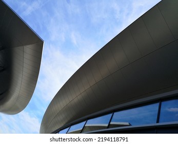 Modern Buildings, View From Below. Abstract Architecture And Material Design With Curvy Hitech Roof. Minimal Exterior Background With Curve Structure. Geometric Composition Of Curves Against Blue Sky.