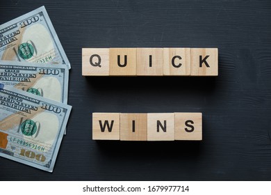 Modern Business Buzzword - Quick Wins. Top View On A Black Board With Dollars And Wooden Blocks. Close Up. Top View.