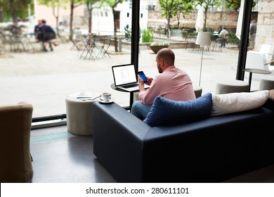 Modern Business Man Busy Working On Smart Phone And Laptop Computer, Young Freelancer Chatting On Cell Phone While Sitting At Sofa Of Coffee Shop, Entrepreneur Using Technology In Hotel Interior