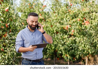 Modern Business, Man Owner Work In Eco Farm, New Normal, Mobile Tech For Growing Organic Fruits. Glad Millennial Handsome Man Typing On Tablet, Talking By Phone, Checking Harvest In Apples Garden