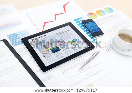 Modern business workplace with business news website on digital tablet, mobile banking on a smartphone and some charts and graphs on a desktop.
