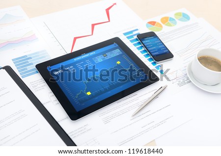 Modern business workplace with stock market data application on a digital tablet, mobile banking interface on a smartphone and some papers with charts, graphs and numbers on a desktop.