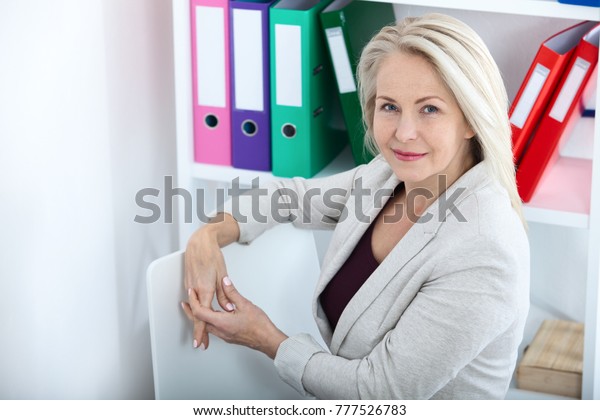 Modern businesswoman. Beautiful middle aged\
woman looking at camera with smile while siting in the office.\
Female face close-up