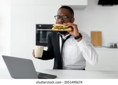 Modern Busy Lifestyle. Portrait Of African Male In Eyeglasses Having Breakfast In Kitchen At Home, Wearing Suit And Drinking Coffee While Eating Sandwich Using Pc Looking At Screen. A Lot Of Work