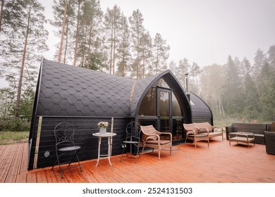 A modern cabin with a black curved roof, wooden deck, and outdoor seating, surrounded by tall trees in a peaceful forest setting. - Powered by Shutterstock