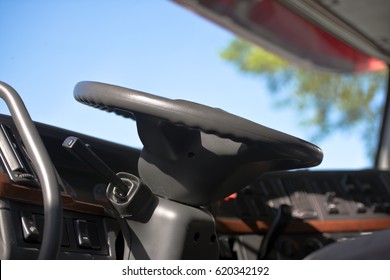 Modern Cabin Of Semi Truck With The Steering Wheel, Column, Dashboard, Control Levers On The Background Of The Windshield Through Which You Can See The Branches Of Trees And Sky