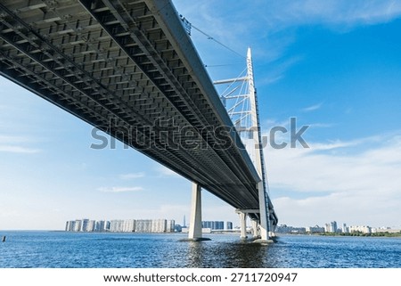 Modern cable stayed bridge from low angle, sweeping over wide blue water with distant Saint Petersburg skyline on the horizon under a clear blue sky. Western High-Speed Diameter (ZSD) bridge
