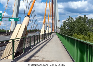 A modern cable-stayed bridge features a pedestrian walkway alongside a multi-lane highway, characterized by bright orange pylons and supports. On the captured scene, the path is enclosed by a tall green railing, offering a clear view of the structural design against a backdrop of a partly cloudy blue sky. Robust concrete pillars rise majestically, anchoring the heavy steel cables that fan out across the horizon, creating a rhythmic visual pattern. To the left, a light-colored vehicle cruises smoothly down the road, passing beneath a prominent blank green directional sign. Lush green trees line the right side, contrasting with the engineered gray asphalt and metallic textures. The perspective emphasizes the linear journey forward, with long, distinct shadows stretching across the pavement, suggesting a bright, sunny day. Engineering precision defines the architecture, blending utility with an aesthetic sense of industrial grandeur. Each element, from the sturdy mesh fencing to the tapered orange masts, serves a specific purpose in supporting the massive span. The atmosphere feels calm and orderly, typical of a well-maintained transport corridor. Pedestrians traversing this area are kept safely separated from high-speed transit by the utilitarian yet striking green barricade. Overhead, the expansive firmament provides a vibrant contrast to the stark, geometric lines of the crossing. This setting captures a blend of urban infrastructure and natural beauty, showcasing how human design integrates with the surrounding landscape to facilitate movement and connectivity. The composition highlights the vast scale of the structure, drawing the eye toward the distant point where the road and the sky converge.