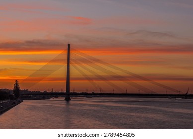 Modern cable-stayed bridge spanning across river at golden sunset, with dramatic orange sky and silhouetted cityscape in background.