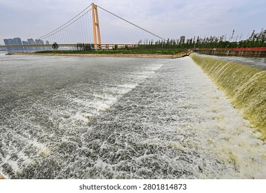 Modern cable-stayed bridge spanning a river with active spillway and city skyline, featuring curved architectural design and urban landscape infrastructure.