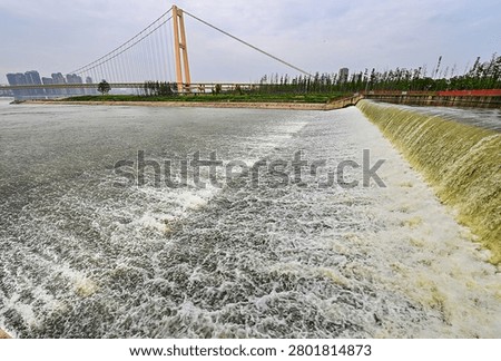 Modern cable-stayed bridge spanning a river with active spillway and city skyline, featuring curved architectural design and urban landscape infrastructure.