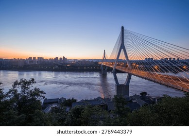 Modern cable-stayed bridge spanning wide river at sunrise with city skyline illuminated in golden light and calm water reflections below.