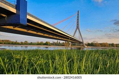 A modern cable-stayed bridge spans a tranquil river with lush green grass and a clear blue sky in the background.