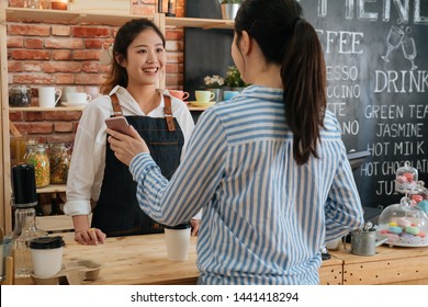 In Modern Cafe Friendly Woman Worker Make Takeaway Coffee For Office Lady Customer Who Pay By Contactless Mobile Phone To Credit Card System. Client Showing Online Payment On Smartphone To Waitress