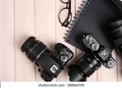 Modern cameras with glasses and notebook on wooden table, top view