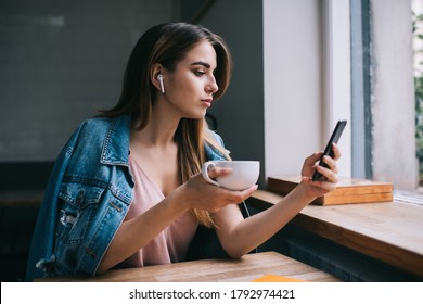 Modern casual female in denim drinking coffee while surfing Internet on smartphone with wireless earbuds spending free time in urban cafe
