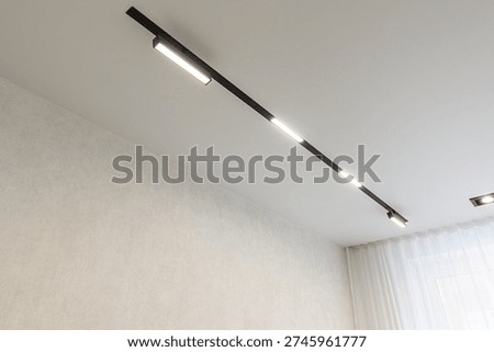 A modern ceiling features a sleek black linear track lighting system with three integrated LED modules; light-colored textured walls and sheer white curtains in the background.