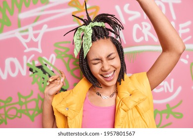 Modern cheerful girl with trendy hairstyle has upbeat mood dances carefree keeps arms raised up laughs positively poses against graffiti wall dressed in stylish clothes. Street rock style concept - Shutterstock ID 1957801567