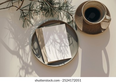 Modern Christmas stationery. Blank greeting card, invitation. Envelope mockup in sunlight on plate. Green pine tree branches on white table background, long shadows. Cup of coffee, winter flatlay, top - Powered by Shutterstock