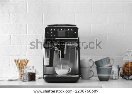 Modern coffee machine pouring milk into glass cup on white countertop in kitchen
