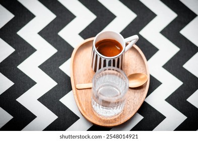 Modern coffee presentation with a striped espresso cup, glass of water, and golden spoon on a wooden tray, placed on a bold black-and-white chevron-patterned background. - Powered by Shutterstock